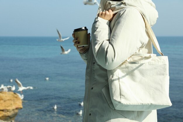 Mujer en chaqueta con vaso de papel y elegante bolsa ecológica contra el mar