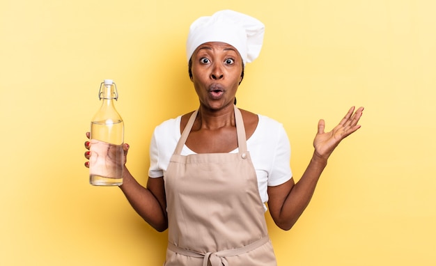 Mujer chef afro negra con la boca abierta y asombrada, conmocionada y asombrada con una increíble sorpresa sosteniendo una botella de agua