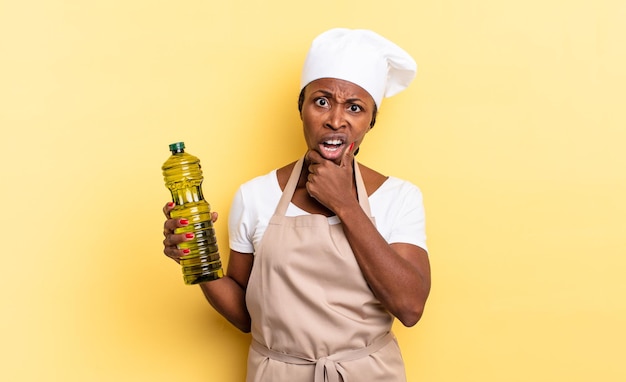 Mujer chef afro negra con la boca y los ojos bien abiertos y la mano en la barbilla, sintiéndose desagradablemente conmocionada, diciendo qué o guau. concepto de aceite de oliva