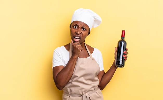 Mujer chef afro negra con la boca y los ojos bien abiertos y la mano en la barbilla sintiéndose desagradablemente sorprendida diciendo qué o wow concepto de botella de vino