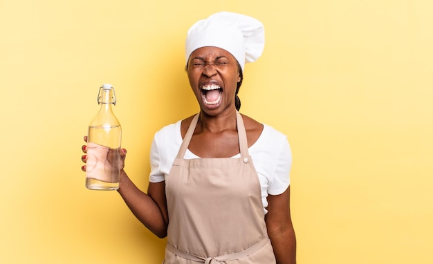 Mujer chef afro negra gritando agresivamente luciendo muy enojada frustrada indignada o molesta gritando sin sostener una botella de agua