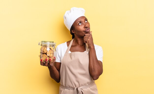 Mujer chef afro negra pensando, sintiéndose dudosa y confundida, con diferentes opciones, preguntándose qué decisión tomar. concepto de cookies