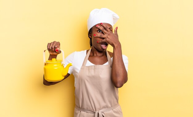 Mujer chef afro negra que parece sorprendida, asustada o aterrorizada, cubriendo la cara con la mano y mirando entre los dedos. concepto de tetera