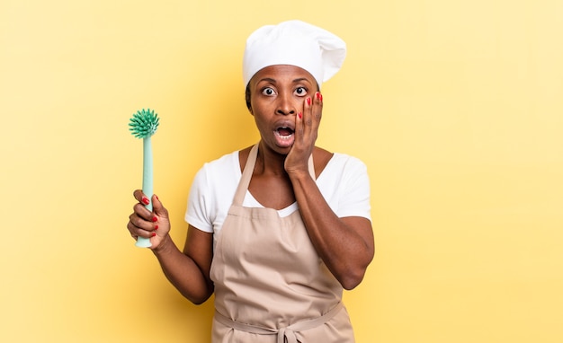 Mujer de chef afro negra que se siente conmocionada y asustada, mirando aterrorizada con la boca abierta y las manos en las mejillas. concepto de limpieza de platos