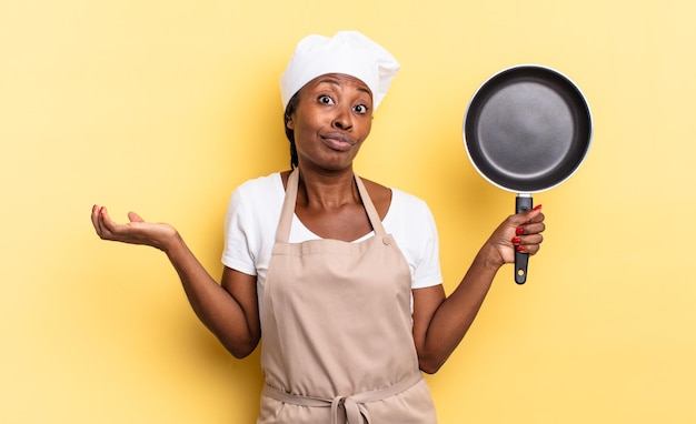 Mujer chef afro negra que se siente desconcertada y confundida, dudando, ponderando o eligiendo diferentes opciones con expresión divertida