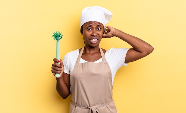 Mujer chef afro negra que se siente estresada, preocupada, ansiosa o asustada, con las manos en la cabeza, en pánico por el error. concepto de limpieza de platos