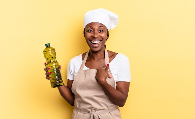 Mujer chef afro negra que se siente feliz, sorprendida y orgullosa, señalando a sí misma con una mirada emocionada y asombrada. concepto de aceite de oliva