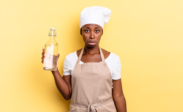 Mujer chef afro negra que se siente triste y quejumbrosa con una mirada infeliz llorando con una actitud negativa y frustrada sosteniendo una botella de agua