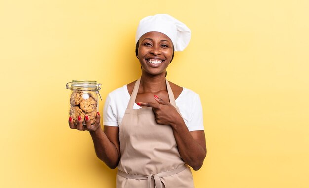 Mujer chef afro negra sonriendo alegremente, sintiéndose feliz y apuntando hacia un lado y hacia arriba, mostrando objetos en el espacio de la copia. concepto de galletas