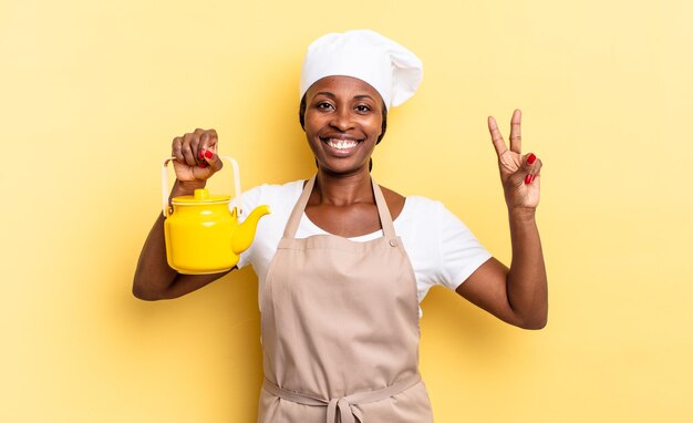 Mujer chef afro negra sonriendo y luciendo amigable, mostrando el número dos o segundo con la mano hacia adelante, contando hacia atrás. concepto de tetera