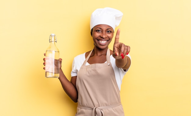 Mujer chef afro negra sonriendo con orgullo y confianza haciendo la pose número uno triunfalmente, sintiéndose como un líder sosteniendo una botella de agua