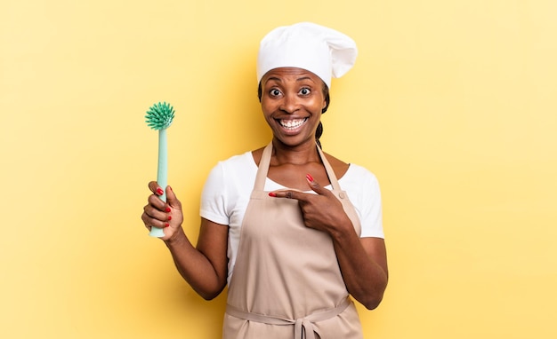 Mujer de chef afro negro mirando emocionada y sorprendida apuntando hacia un lado y hacia arriba para copiar el espacio. concepto de limpieza de platos