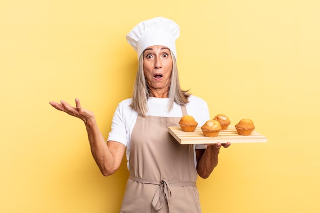 Mujer chef de mediana edad con la boca abierta y asombrada, conmocionada y asombrada con una sorpresa increíble y sosteniendo una bandeja de muffins