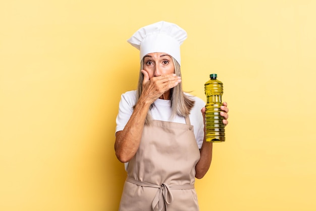 Mujer chef de mediana edad cubriendo la boca con las manos con una expresión de sorpresa y sorpresa, manteniendo un secreto o diciendo ¡uy!