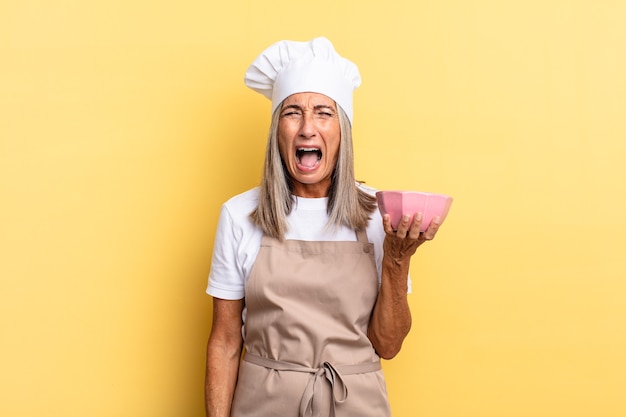 Mujer chef de mediana edad gritando agresivamente, luciendo muy enojada, frustrada, indignada o molesta, gritando no y sosteniendo una olla vacía