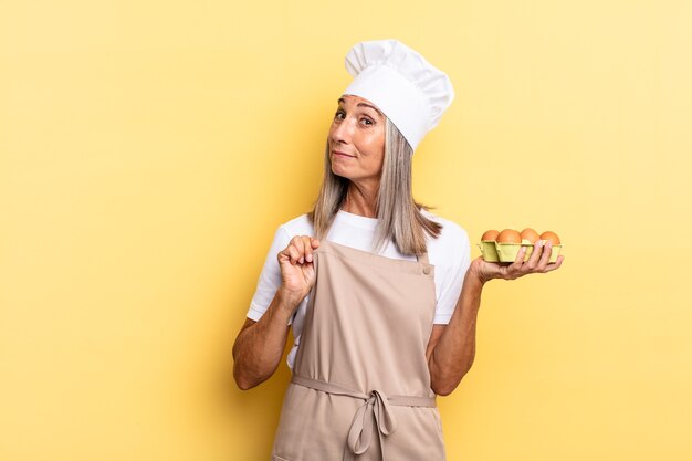 Mujer chef de mediana edad que parece arrogante, exitosa, positiva y orgullosa, apuntando a sí mismo sosteniendo una caja de huevos