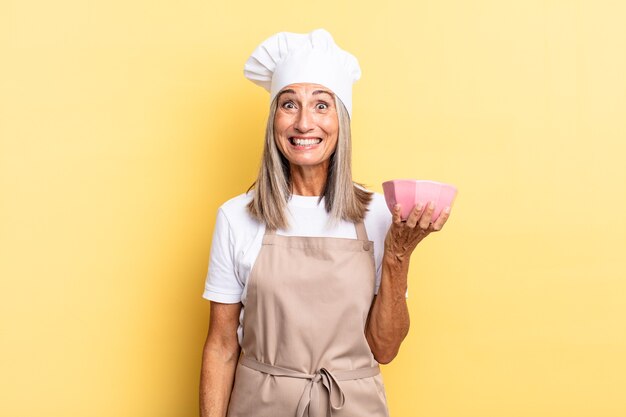 Mujer chef de mediana edad que parece feliz y gratamente sorprendida, emocionada con una expresión fascinada y conmocionada y sosteniendo una olla vacía