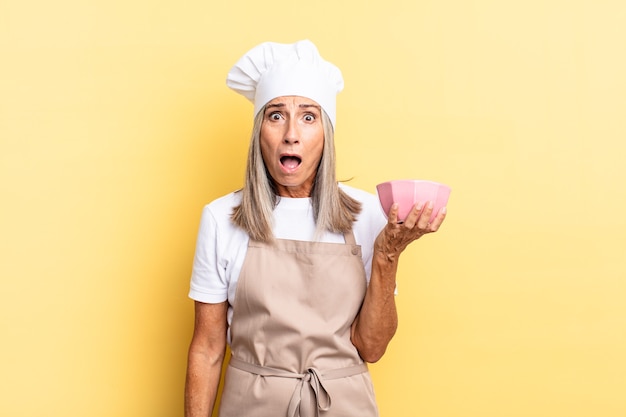 Mujer chef de mediana edad que parece muy conmocionada o sorprendida, mirando con la boca abierta diciendo wow y sosteniendo una olla vacía