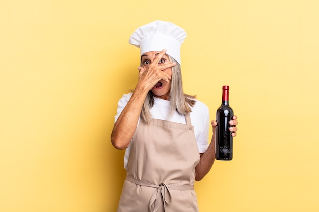 Mujer chef de mediana edad que parece sorprendida, asustada o aterrorizada, cubriendo la cara con la mano y mirando entre los dedos sosteniendo una botella de vino