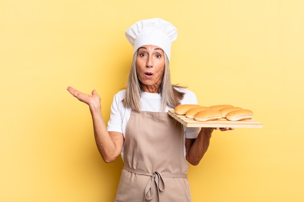 Mujer chef de mediana edad que parece sorprendida y conmocionada, con la mandíbula caída sosteniendo un objeto con una mano abierta en el costado y sosteniendo una bandeja de pan