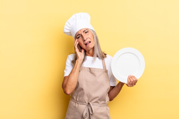 Mujer chef de mediana edad que se siente aburrida, frustrada y con sueño después de una tarea tediosa, aburrida y tediosa, sosteniendo la cara con la mano y sosteniendo un plato