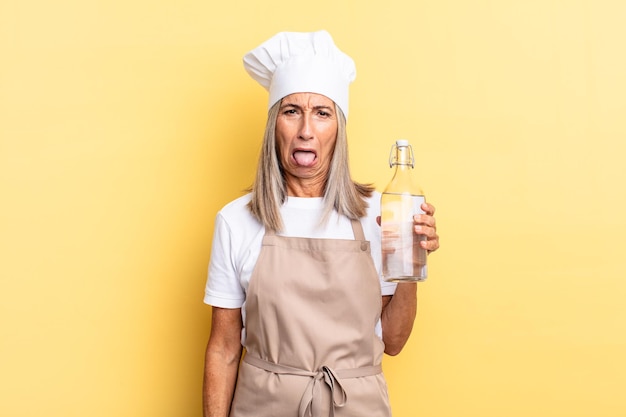 Mujer chef de mediana edad que se siente disgustada e irritada, sacando la lengua, disgustando algo desagradable y asqueroso con una botella de agua