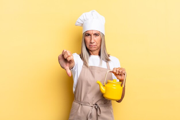 Mujer chef de mediana edad que se siente enojada, molesta, decepcionada o disgustada, mostrando los pulgares hacia abajo con una mirada seria y sosteniendo una tetera