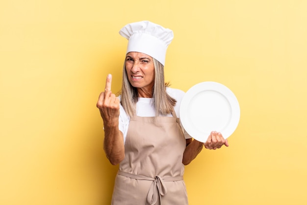 Mujer chef de mediana edad que se siente enojada, molesta, rebelde y agresiva, moviendo el dedo medio, contraatacando y sosteniendo un plato