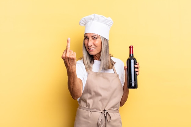 Mujer chef de mediana edad que se siente enojada, molesta, rebelde y agresiva, moviendo el dedo medio, luchando sosteniendo una botella de vino