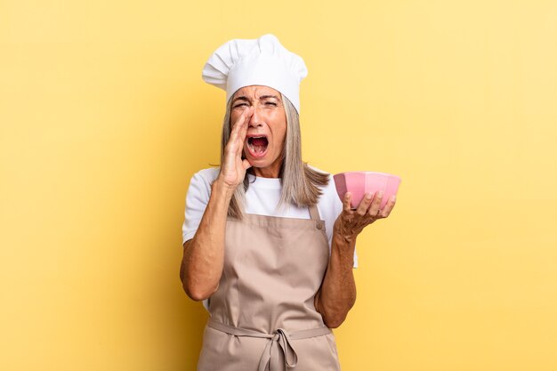 Mujer chef de mediana edad que se siente feliz, emocionada y positiva, dando un gran grito con las manos al lado de la boca, gritando y sosteniendo una olla vacía