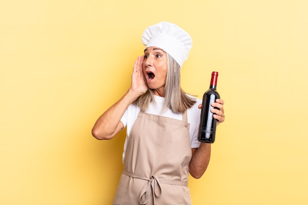 Mujer chef de mediana edad que se siente feliz, emocionada y sorprendida, mirando hacia el lado con ambas manos en la cara sosteniendo una botella de vino