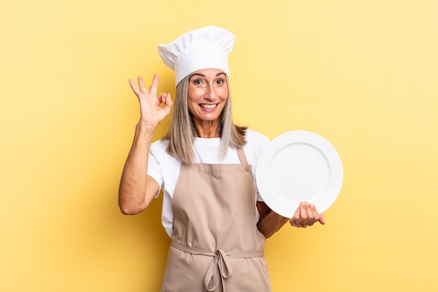 Mujer chef de mediana edad que se siente feliz, relajada y satisfecha, mostrando aprobación con un gesto aceptable, sonriendo y sosteniendo un plato