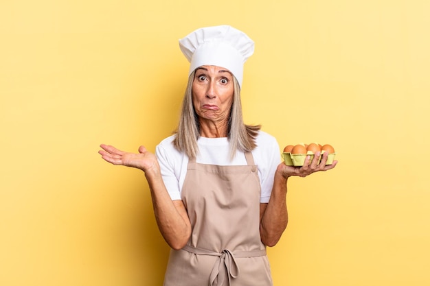 Mujer chef de mediana edad que se siente perpleja y confundida, dudando, ponderando o eligiendo diferentes opciones con expresión divertida sosteniendo una caja de huevos