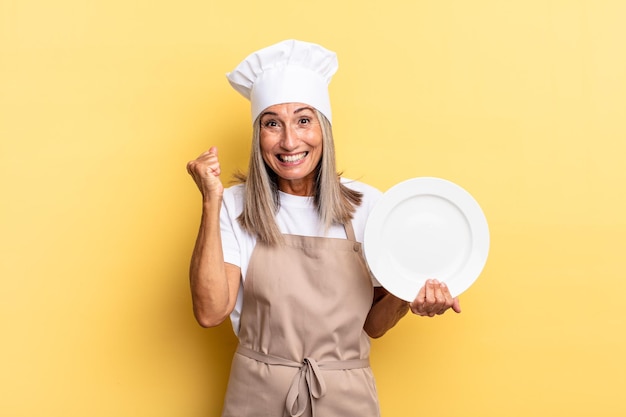 Mujer chef de mediana edad que se siente sorprendida, emocionada y feliz, riendo y celebrando el éxito, diciendo ¡guau! y sosteniendo un plato