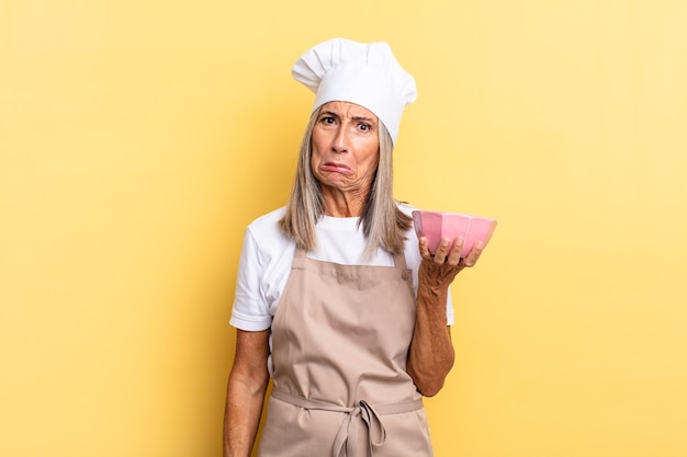 Mujer chef de mediana edad que se siente triste y llorona con una mirada infeliz, llorando con una actitud negativa y frustrada y sosteniendo una olla vacía