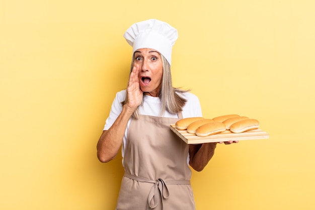 Mujer chef de mediana edad sintiéndose conmocionada y asustada, mirando aterrorizada con la boca abierta y las manos en las mejillas y sosteniendo una bandeja de pan