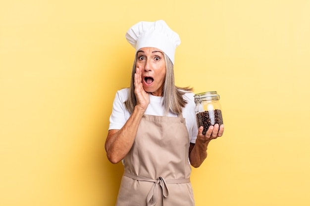 Mujer chef de mediana edad sintiéndose conmocionada y asustada, mirando aterrorizada con la boca abierta y las manos en las mejillas sosteniendo granos de café