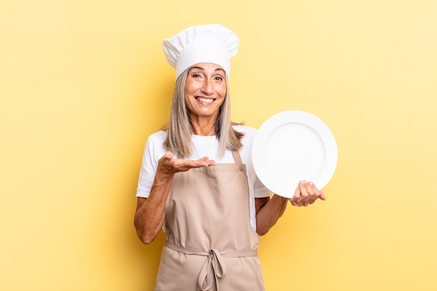 Mujer chef de mediana edad sonriendo alegremente, sintiéndose feliz y mostrando un concepto en el espacio de la copia con la palma de la mano y sosteniendo un plato