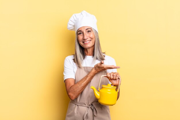 Mujer chef de mediana edad sonriendo alegremente, sintiéndose feliz y mostrando un concepto en el espacio de la copia con la palma de la mano y sosteniendo una tetera