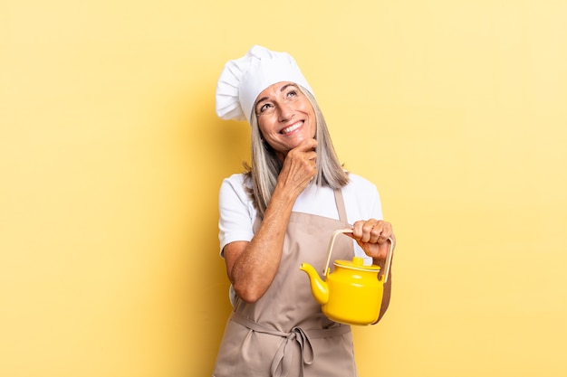 Mujer chef de mediana edad sonriendo con una expresión feliz y segura con la mano en la barbilla, preguntándose y mirando hacia un lado y sosteniendo una tetera