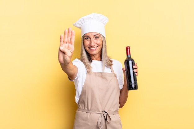 Mujer chef de mediana edad sonriendo y mirando amistosamente, mostrando el número cuatro o cuarto con la mano hacia adelante, contando hacia atrás sosteniendo una botella de vino