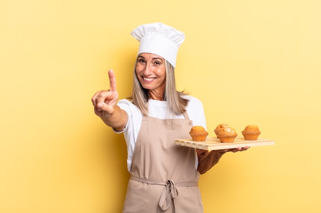 Mujer chef de mediana edad sonriendo y mirando amistosamente, mostrando el número uno o el primero con la mano hacia adelante, contando hacia atrás y sosteniendo una bandeja de muffins