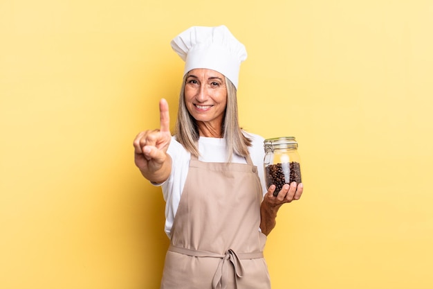 Mujer chef de mediana edad sonriendo y mirando amistosamente, mostrando el número uno o el primero con la mano hacia adelante, contando hacia atrás sosteniendo los granos de café