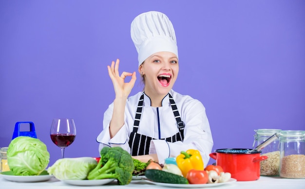 Mujer chef usa sombrero delantal cerca de los ingredientes de la mesa Chica adorable chef enseña culinaria Las mejores recetas culinarias para probar en casa Receta perfecta Convierte los ingredientes en una deliciosa comida Habilidades culinarias