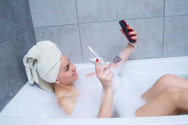 Mujer chica con una toalla en la cabeza se acuesta con una copa de champán en una bañera blanca y toma una selfie en el teléfono. En sus manos mucho jabón borracho. Relájate después de un día duro. Procedimiento de relajación del spa