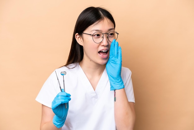 Mujer china dentista sosteniendo herramientas aisladas en un fondo beige gritando con la boca abierta a un lado