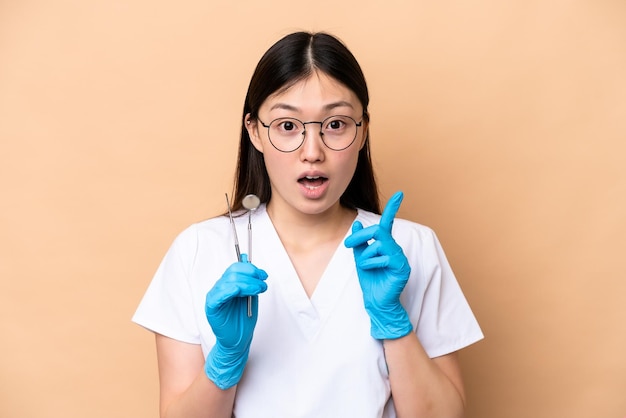 Mujer china dentista sosteniendo herramientas aisladas en un fondo beige con la intención de darse cuenta de la solución mientras levanta un dedo