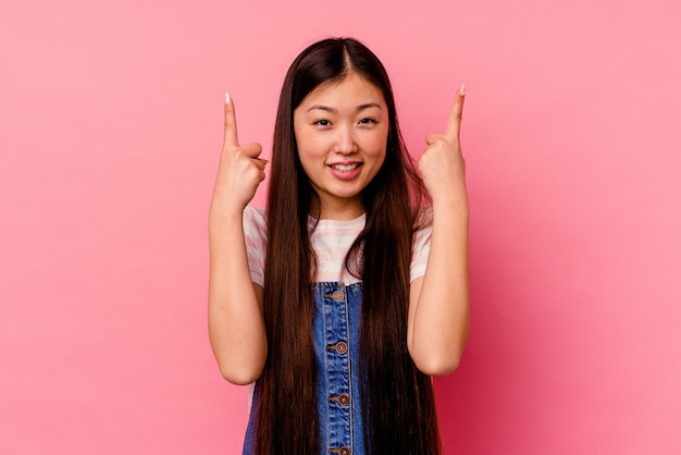 La mujer china joven aislada en la pared rosada indica con ambos dedos índice hacia arriba mostrando un espacio en blanco.