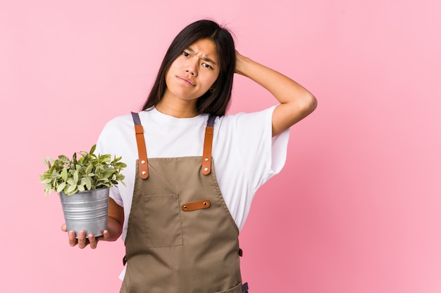 La mujer china joven del jardinero que sostenía una planta aisló tocar la parte posterior de la cabeza, pensando y tomando una decisión.