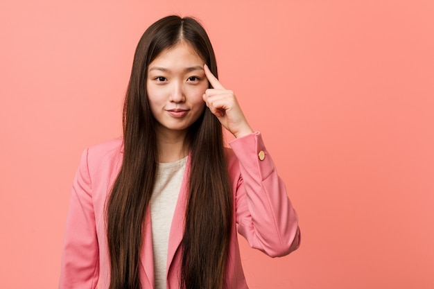 Mujer china joven del negocio que lleva el templo rosado que señala el templo con el dedo, pensando, centrado en una tarea.